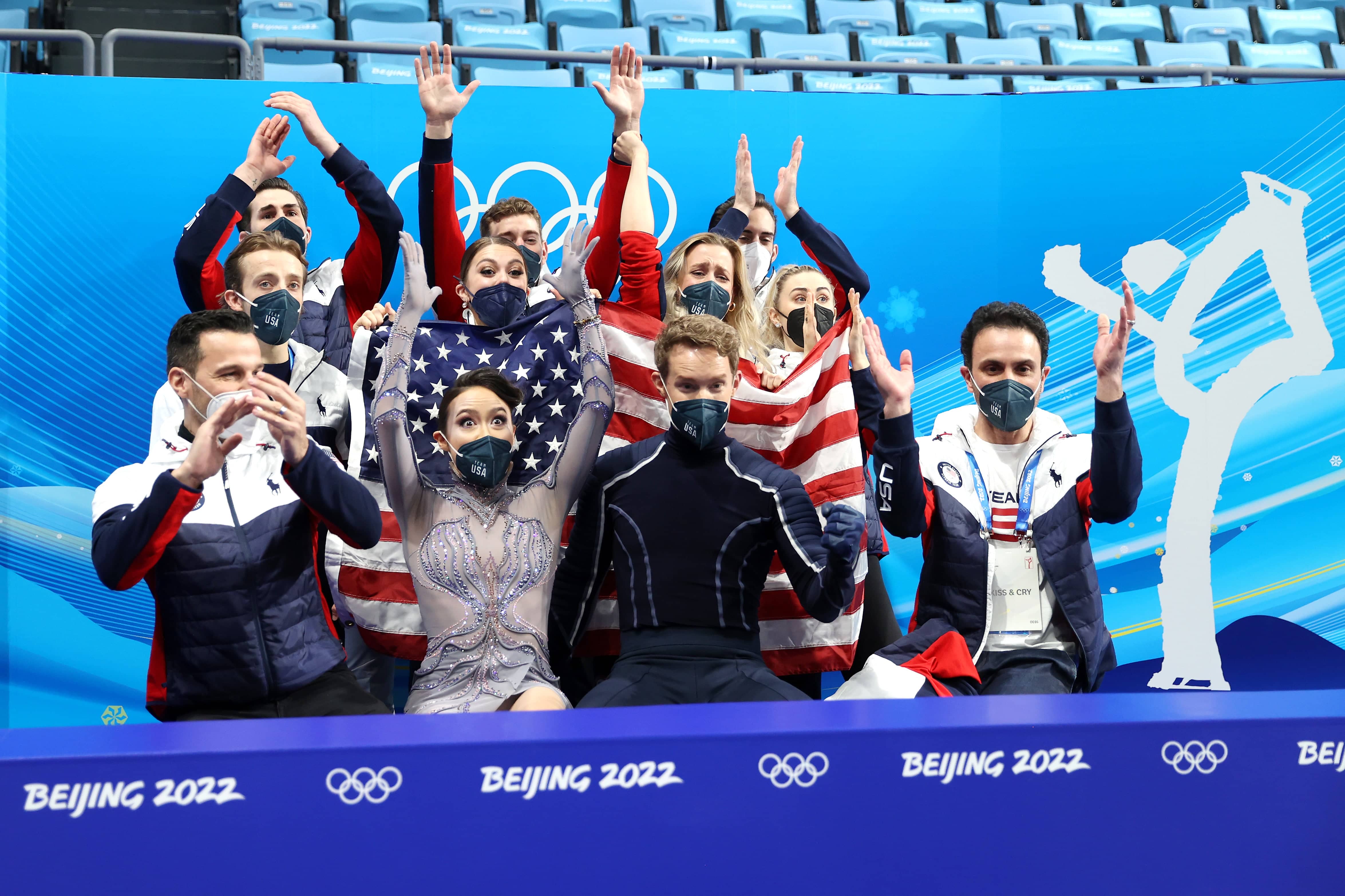 Taem USA, in their box, celebrates Chock and Bates' score, all athletes and coaches with their arms raised and waving the USA Flag